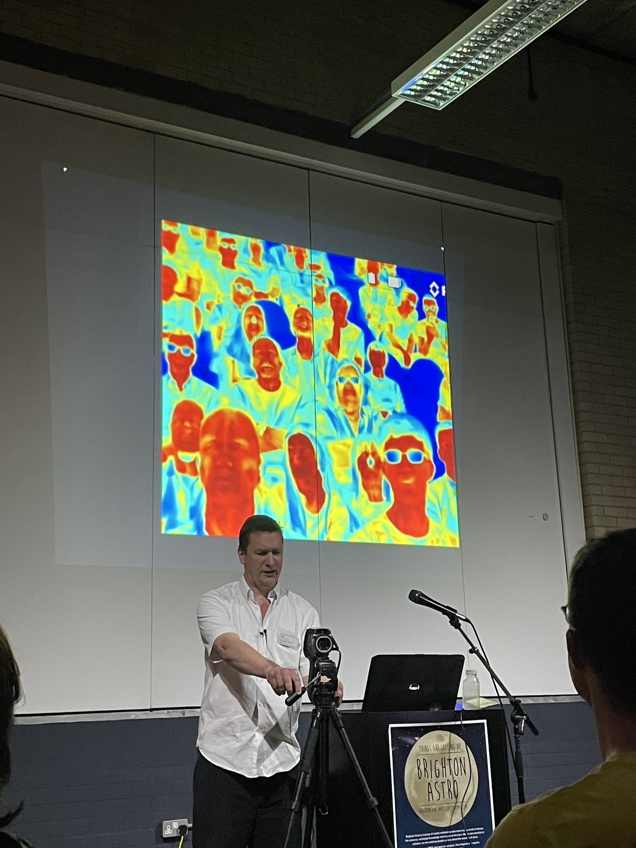 the speaker behind an infrared camera, pointed at the audience, with the image projected showing faces in red and bodies in greens and blues. glasses appear dark.