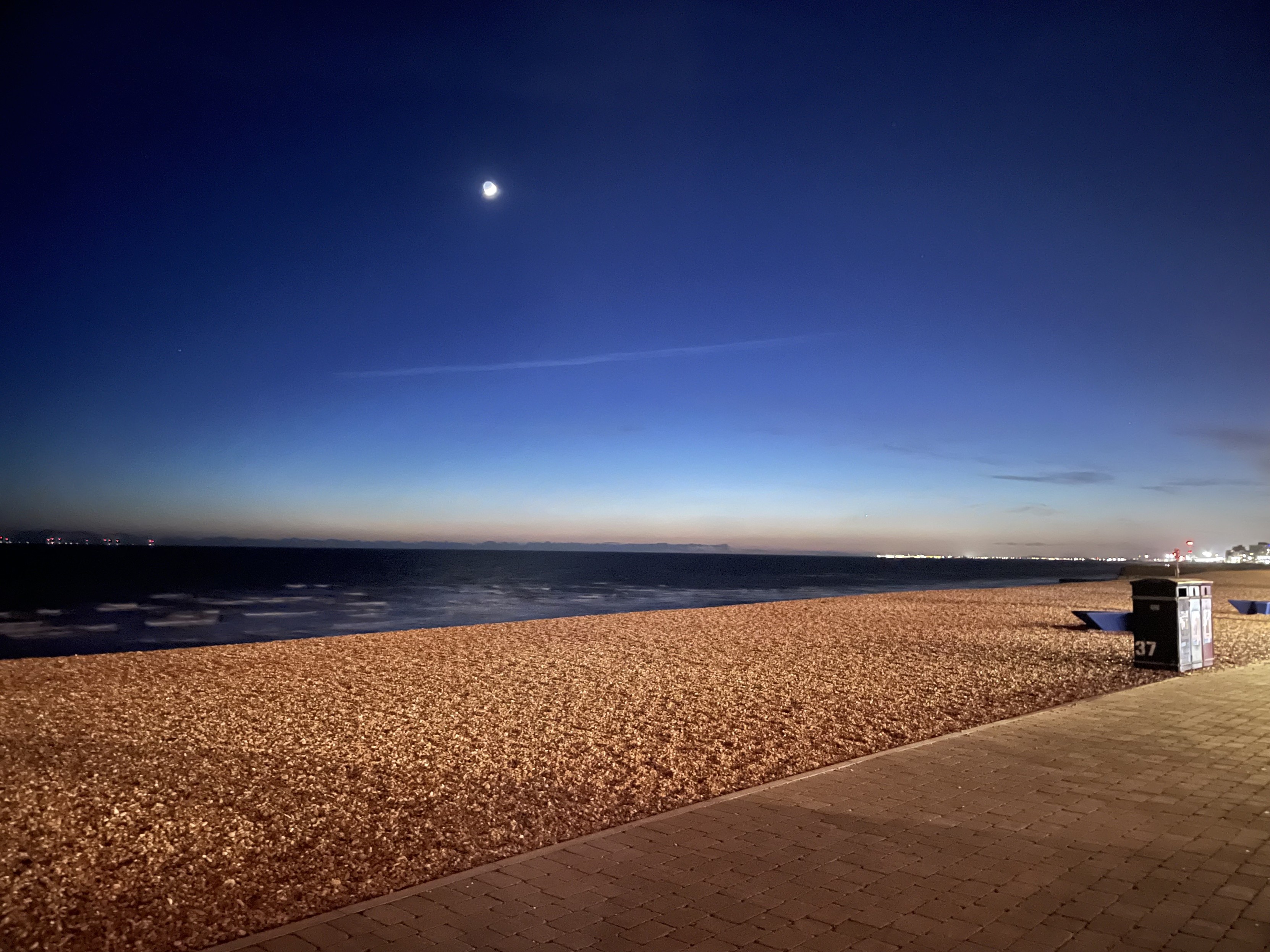 An early evening view of Brighton Beach, looking out over the pebbles with three bins on the right edge of the image, and a thin crescent moon in the sky.