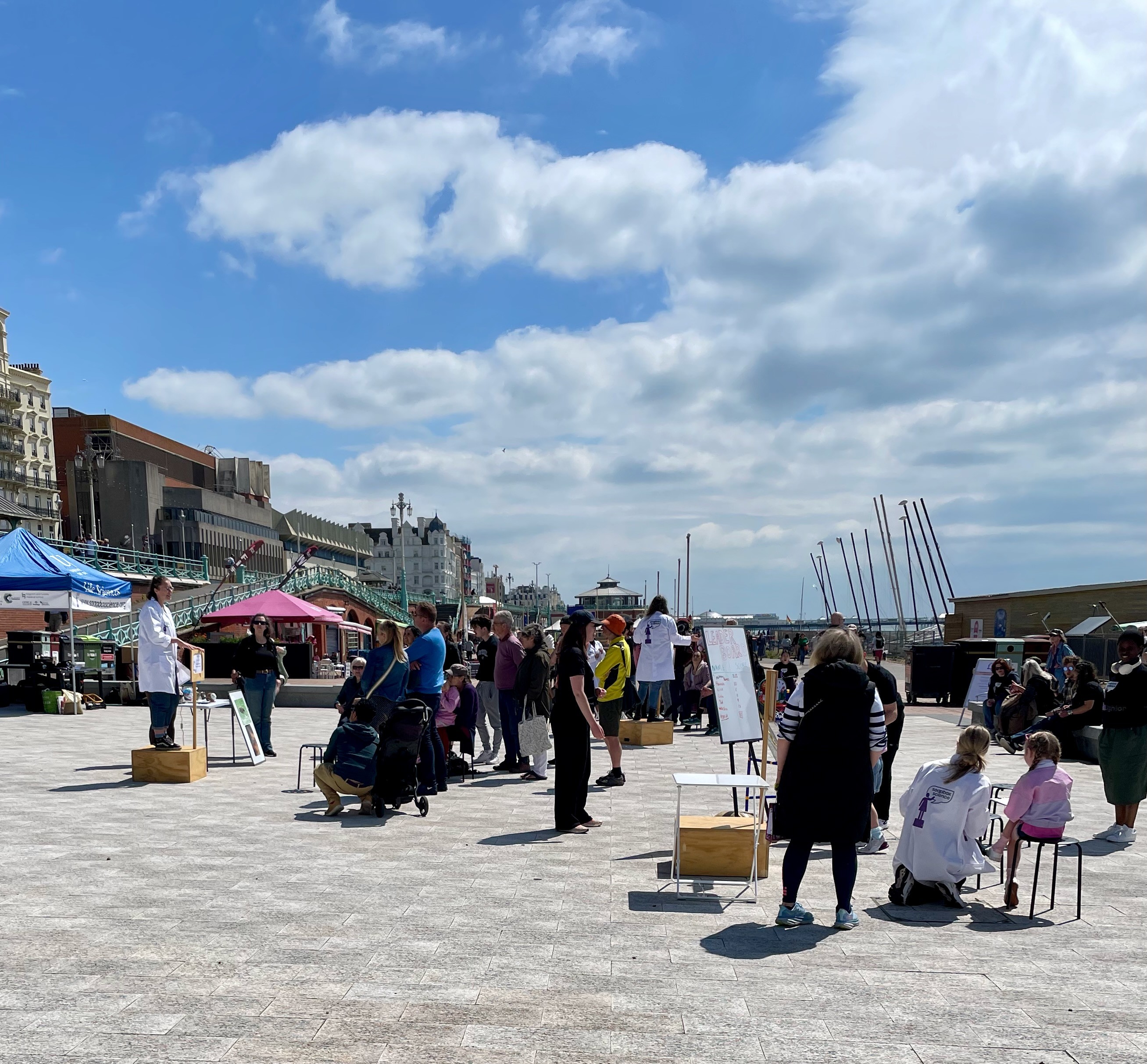 View onto the square on the Brighton seafront where soapbox science is held. Three boxes visible, where a piece of science is being discussed at each one with a small crowd around. Blue skies, and fluffy white clouds.